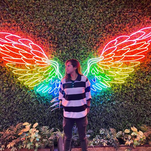 Une personne se tient debout entre deux ailes néon au couleurs de l'arc-en-ciel.